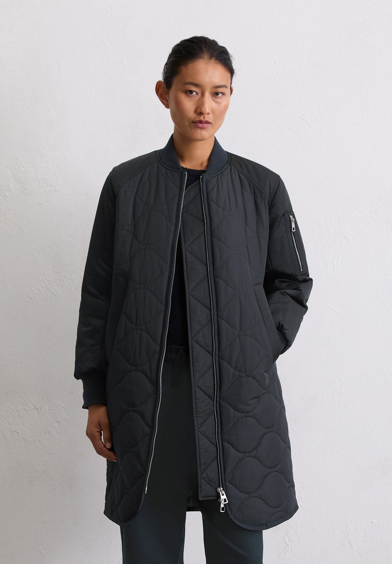 Femme aux cheveux bruns portant une longue veste matelassée noire avec fermeture éclair à l'avant, col et poignets côtelés, et poche à fermeture éclair sur la manche, se tenant devant un mur blanc.