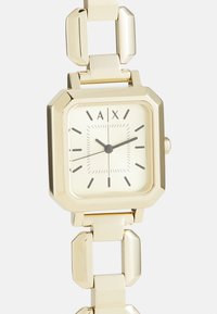 Montre-bracelet carrée ton or avec cadran crème, index heures noirs minimalistes et bracelet en chaîne métallique reliée montrant l'heure 2:10:40.