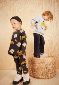 Zwei Kinder tragen spielerische Outfits: eines in einem dunklen, bedruckten Oberteil und Leggings mit Lebensmittel-Icons; das andere in einem grauen Sweatshirt und Cordhosen.