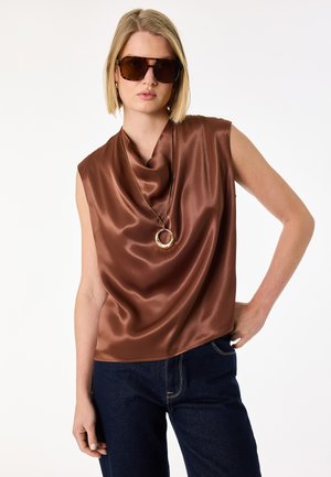 Blondynka w brązowej satynowej bluzce bez rękawów, ciemnych okularach przeciwsłonecznych, złotym naszyjniku kołach i ciemnoniebieskich dżinsach, stojąca z jedną ręką na biodrze.