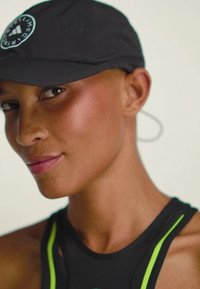 Sort kasket med et cirkulært logo, båret på skrå. Personen har også en sort atletisk top med grønne detaljer, som fremviser en glat hudstruktur.