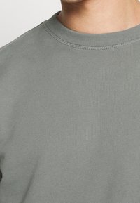 Zbliżenie na osobę noszącą jednolitą szaro-skalową bluzę z okrągłym dekoltem, ukazującą linię dekoltu i górną część klatki piersiowej.