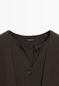 Cardigan marron foncé avec une texture lisse, un col rond et trois boutons le long de la patte de boutonnage, avec une étiquette de marque visible.