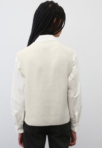 Cremfarbener ärmelloser Strickpullover mit strukturiertem Finish, ausgestattet mit einem gerippten Saum und einem schlichten Design, getragen über einem weißen Hemd mit Knöpfen.