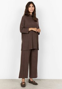 Tunic in maglia marrone con collo alto e maniche a tre quarti, abbinata a pantaloni larghi coordinati e scarpe slip-on con borchie.