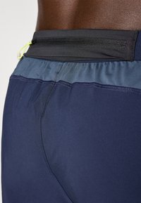 Marinefarbene Sportshorts mit einem schwarzen Bund, der eine Reißverschlusstasche und elastische Details aufweist. Glattes, leichtes Material mit einem schlanken Design.