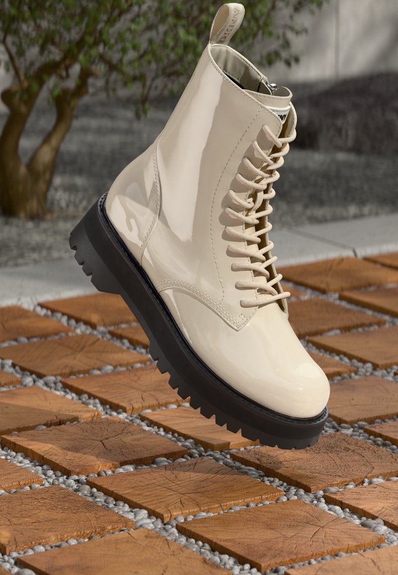 Botas de tobillo de charol beige con diseño de cordones, suela de goma negra texturizada y tirador en la parte superior, exhibidas sobre una superficie de madera.