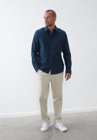 Camisa de botones azul marino con un patrón sutil, combinada con pantalones beige y zapatillas blancas, de pie frente a una pared blanca lisa.