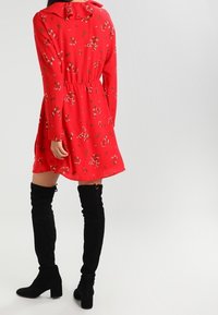Abito floreale rosso con colletto arricciato, vita stretta e maniche lunghe. Abbinato a stivali alti fino alla coscia in suede nero.