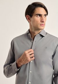 Camisa de botones gris con mangas largas, tejido ligero, textura sutil y pequeño logo bordado en el pecho. Presenta puños estándar y botones.