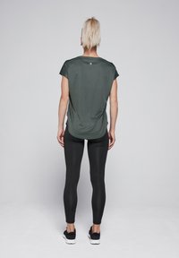 Dark green athletic shirt with short sleeves, relaxed fit, and rounded hem; paired with black leggings and black athletic shoes.