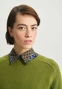 Pull en maille verte avec un tissu texturé et un col ras du cou, superposé sur une chemise à col noire et bleue à motifs floraux.