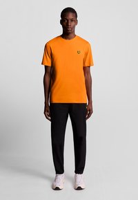 T-shirt di cotone arancione con collo a giro e maniche corte, che presenta un piccolo logo nero in evidenza. Abbinato a pantaloni neri e sneakers chiare.