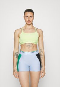 Svijetlo limeta zelena sportska grudnjak s tankim trakama i malim adidas logom. Uparen s svijetloplavim šortsevima koji imaju zelene i crne bočne panele.