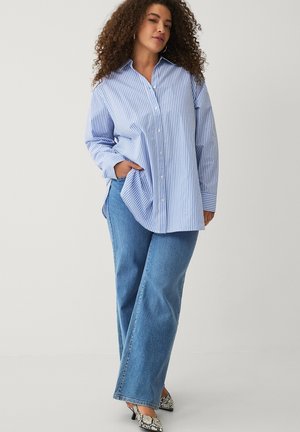 Frau mit lockigem Haar, die ein übergroßes blau-weiß gestreiftes Hemd, blaue Jeans und High Heels mit Schlangenmuster trägt und mit einer Hand in der Tasche posiert.