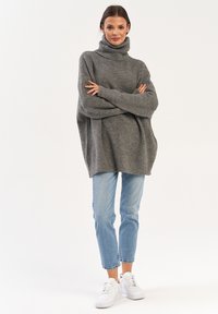 Szary, oversize sweter z golfem o prążkowanej strukturze, zestawiony z jasnoniebieskimi spodniami prostymi i białymi trampkami, stojący na jednolitym tle.