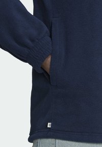 Marinblå fleecetröja med elastiska muddar, sidofickor och en liten logotypflik vid fållen, med en mjuk, texturerad yta.