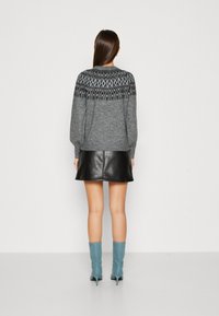 Femme debout, portant un sweater à motif gris, une jupe en cuir noire et des bottines à talons bleues, vue de dos sur un fond uni.