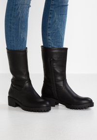 Botas de cuero negras hasta la rodilla con un acabado texturizado, cremallera lateral y suela de goma gruesa. Combinadas con unos jeans de mezclilla azul ajustados.