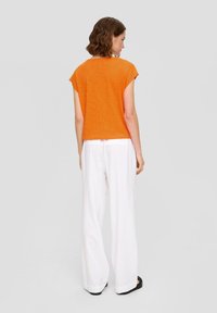 Oranje top met korte mouwen, gecombineerd met witte wijde broek, van achteraf weergegeven. Top heeft een ontspannen pasvorm; de broek is lichtgewicht en vloeiend.