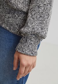 Grå och vit stickad tröja med ribbade ärmar, ihopparad med blå denimjeans. Närbild av en hand som vilar mot tröjans ärm.