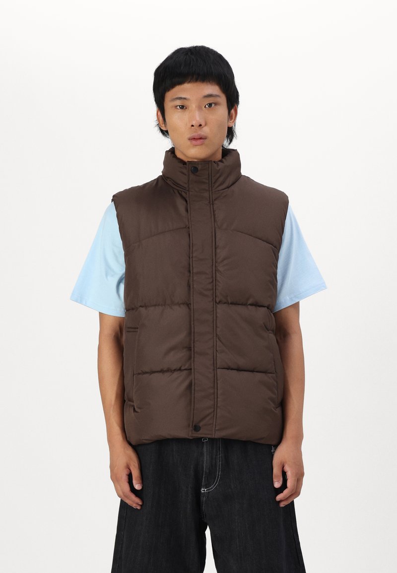 Brown puffer vest with high collar, button closure, and side pockets, paired with a light blue t-shirt. The vest features a quilted texture.