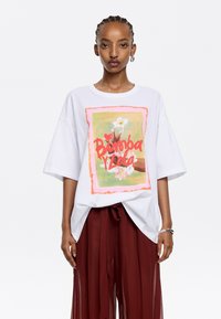 Una camiseta blanca de gran tamaño presenta un gráfico colorido de una mano sosteniendo flores con el texto "Binoba y Lola", sobre un suave fondo verde.