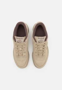Beige mockasin sneakers med rund tå, perforerad detaljering och brun textilfoder. Har snörning och en Reebok-logotyp på tungan.