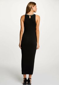 Abito midi nero a coste con dettaglio a chiave sul retro e scollatura decorata. Silhouette aderente che si estende fino alle caviglie. Indossato con sandali neri con tacco.