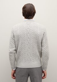 Strickpullover in Hellgrau mit aufwendigen Kabelmustern, Rundhalsausschnitt, dicken Rippbündchen und Saum. Wird über grauen Hosen getragen.