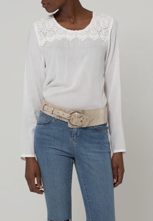 Kobieta w białej bluzce z długim rękawem z kwiatową koronkową linią dekoltu, ubrana w jasnoniebieskie dżinsy i szeroki beżowy teksturowany pasek z okrągłą klamrą.