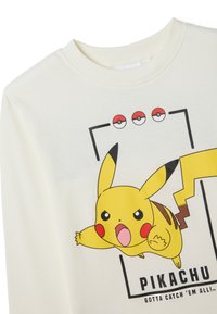 Langärmeliges cremefarbenes T-Shirt mit einem lebhaften gelben Pikachu-Graphic, umrandet von schwarzen Konturen und Text, sowie drei Pokéball-Icons darüber.