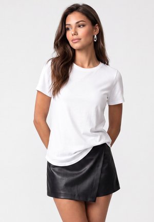 Kvinde med langt brunt hår iført hvid kortærmet t-shirt og sort læder omslagsnederdel, står med hænderne på ryggen og kigger til siden.