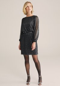 Rochie neagră plisată cu accente metalice, mâneci lungi din material transparent, talie ajustată și tiv volanat. Purtați cu colanți negri transparenți și tocuri argintii.