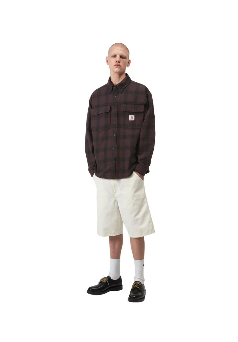 Giovane uomo che indossa una camicia a quadri scura a maniche lunghe, pantaloncini bianchi fino al ginocchio, calzini bianchi e scarpe nere, in piedi con le mani in tasca.