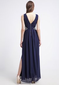 Frau in einem marineblauen, ärmellosen Abendkleid mit tiefem V-Ausschnitt, seitlichem Schlitz, verzierten Taillendetails und nudefarbenen High Heels, stehend vor weißem Hintergrund.