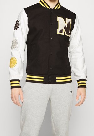 Schwarz-weiße Collegejacke mit gelb gestreiftem Kragen und Bündchen, großem Buchstaben "N" auf der Brust und drei runden Abzeichen am linken Ärmel.