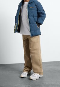 Blaues Puffermantel mit Kapuze, mit kontrastierendem gelbem Reißverschluss und Akzenten. Trägt ein weißes T-Shirt und kombiniert mit beigen Hosen und Turnschuhen.