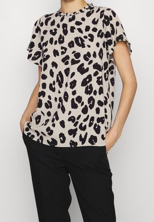 Personne portant un chemisier beige à manches courtes avec des taches léopard noires et un pantalon noir, les mains posées négligemment dans les poches.