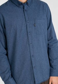 Herrenhemd mit Knöpfen aus dunkelblauem Stoff. Verfügt über eine Tasche auf der linken Brust, kontrastierende Knöpfe und lange Ärmel. Weicher Stoff und lässiges Design.