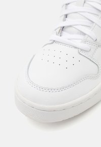 Valkoinen nahkainen urheilusneekeri, jossa on rei'itetty kärki, tasaiset nauhat, teksturoitu kumipohja ja kontrastivärinen ommel reunoilla.