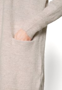Mano nella tasca di un maglione lavorato a maglia beige con polsini a coste su sfondo bianco, mostrando una morbida texture e una vestibilità comoda.