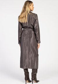 Blonde vrouw in een lange, donkergrijze satijnen jurk met lange mouwen en een ceintuur om de taille, met donkergroefde kniehoge haklaarzen, met de rug naar de kijker gekeerd.