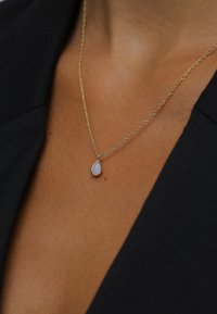 Gouden ketting met een tranenvormige opaal hanger, gezet in een dunne, delicate rand, tegen een textuurrijke huidachtergrond.