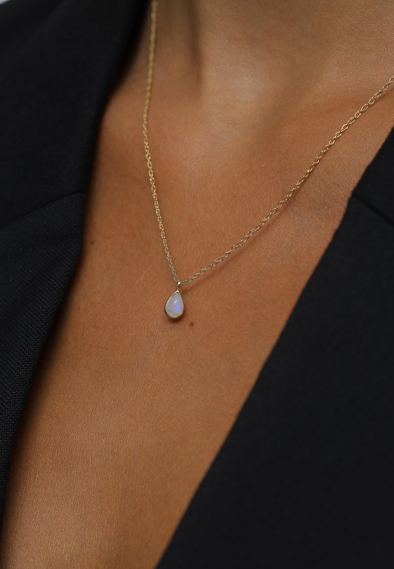 Gouden ketting met een tranenvormige opaal hanger, gezet in een dunne, delicate rand, tegen een textuurrijke huidachtergrond.