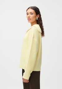 Yellow knitted sweater with a collar, featuring a ribbed texture and loose fit. Paired with dark brown trousers. Side view.