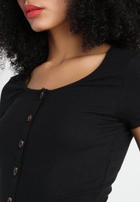 Svart ribbad topp med kort ärm och rundad halsringning, prydd med bruna knappar längs fram för extra detalj.