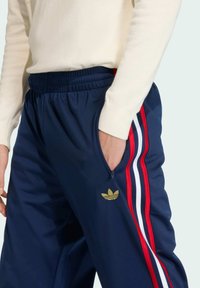 Námornícke jogger nohavice s červenými a bielymi bočnými pruhmi, elastický pás, vrecko na zips, zlaté logo Adidas na ľavom stehne, hladká textúra látky.