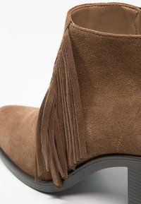 Bottine en daim marron avec un détail frangé sur le côté et un talon bloc en matériau caoutchouteux foncé.