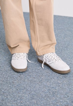 Zapatos de ante en gris con cordones en negro y blanco, con un diseño sencillo y una suela de goma marrón; combinados con pantalones de pierna ancha en beige.